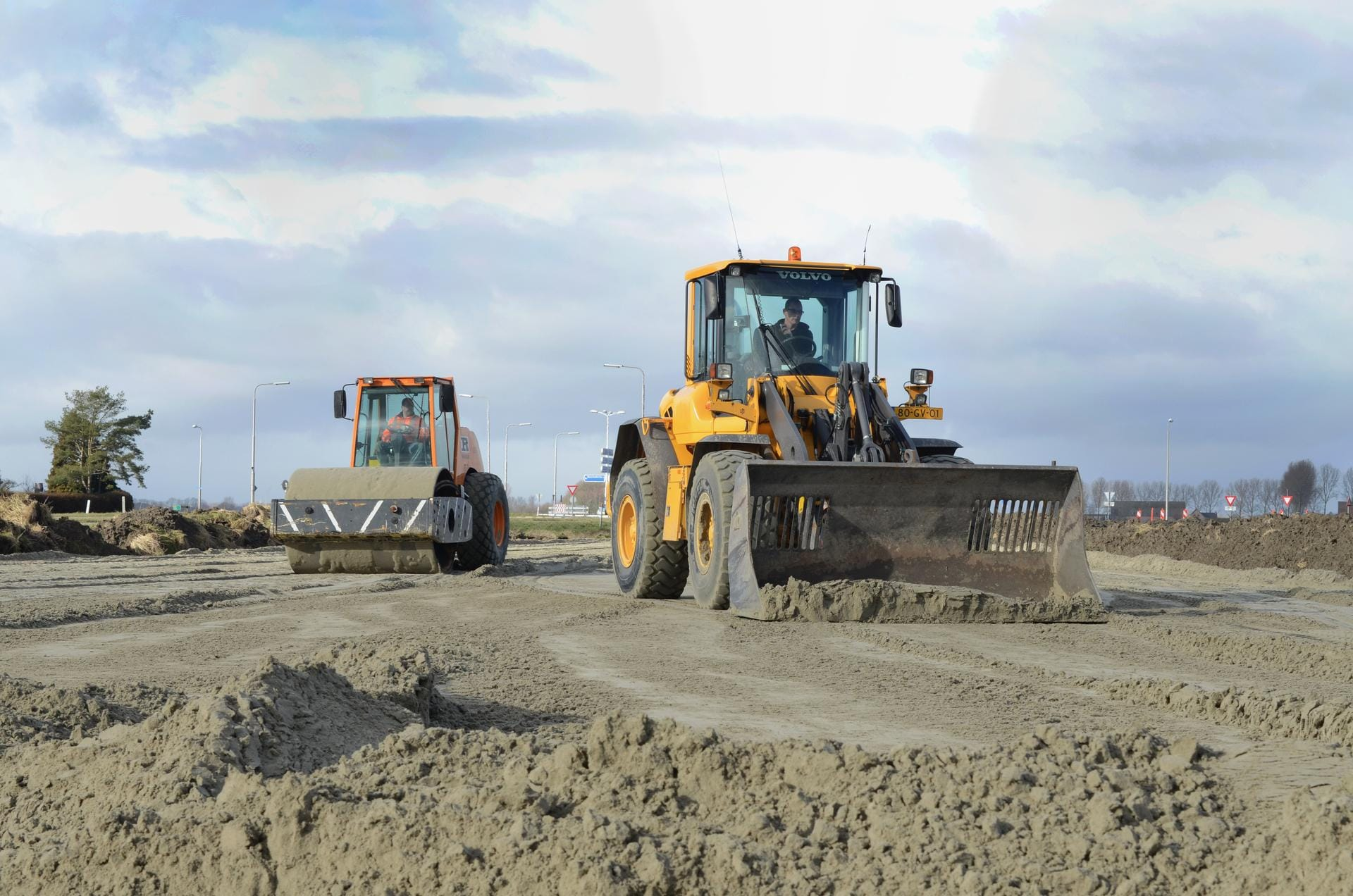 Een shovel die zand schuift op een bouwplaats met daarachter een wals.