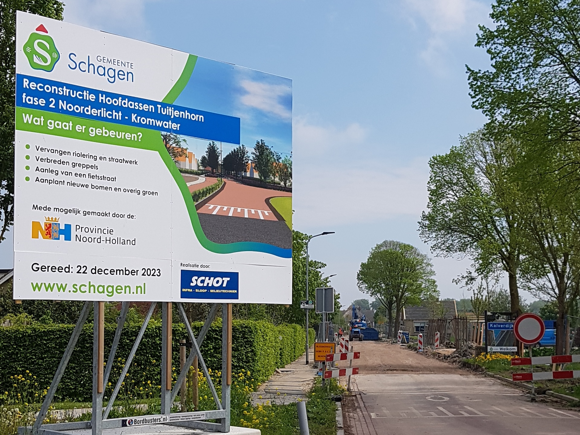 Foto van een bouwbord in Schagen bij een reconstructieproject aan de Hoofdassen in Tuitjenhorn. Het bord vermeldt werkzaamheden zoals riool- en straatwerkvervanging, verbreden greppels, aanleg fietsstraat en aanplant van bomen. Opleverdatum: 22 december 2023. Logo’s van gemeente Schagen, provincie Noord-Holland en aannemer Schot. Op de achtergrond is een afgesloten weg te zien met bouwhekken, borden en graafmachines.