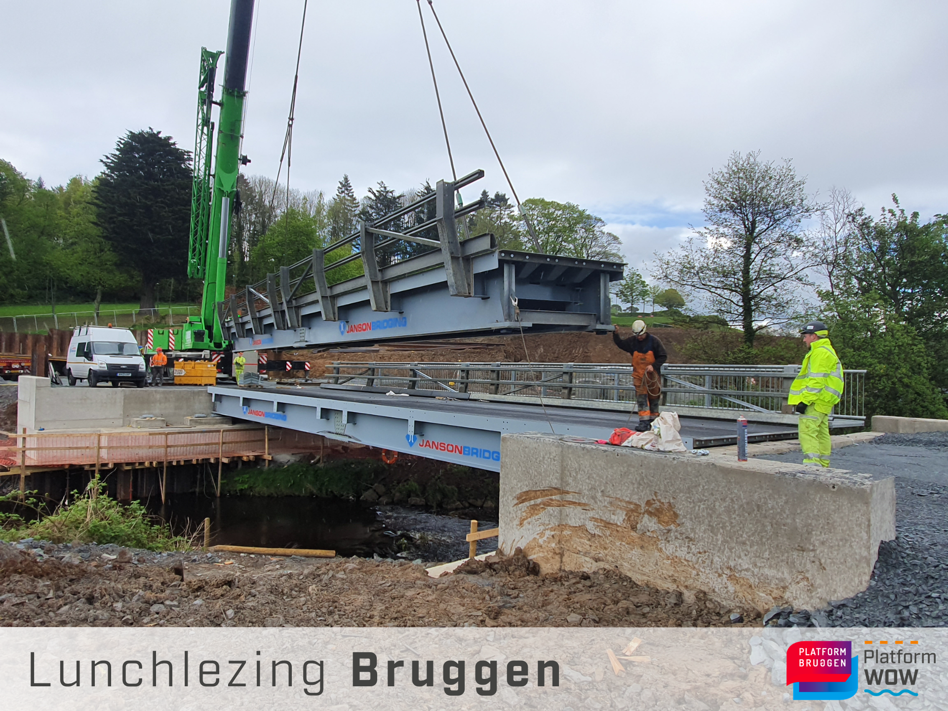 Een brugdeel wordt geplaatst over een beek met een kraan onder begeleiding van 2 bouwvakkers in werkkleding.