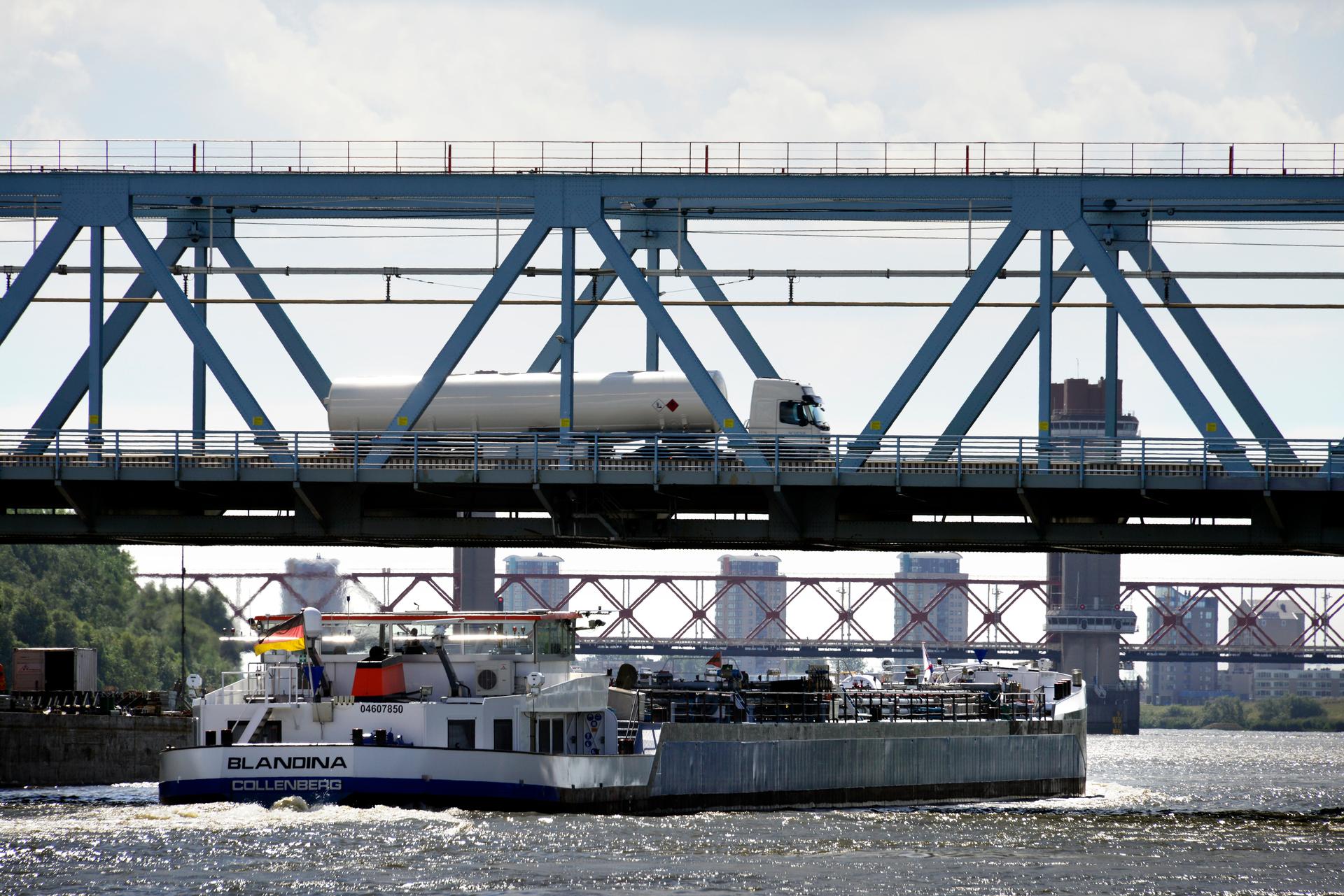 Een brug over het water met op de brug een vrachtwagen en onder de brug een boot. Op de achtergrond de skyline van een stad.