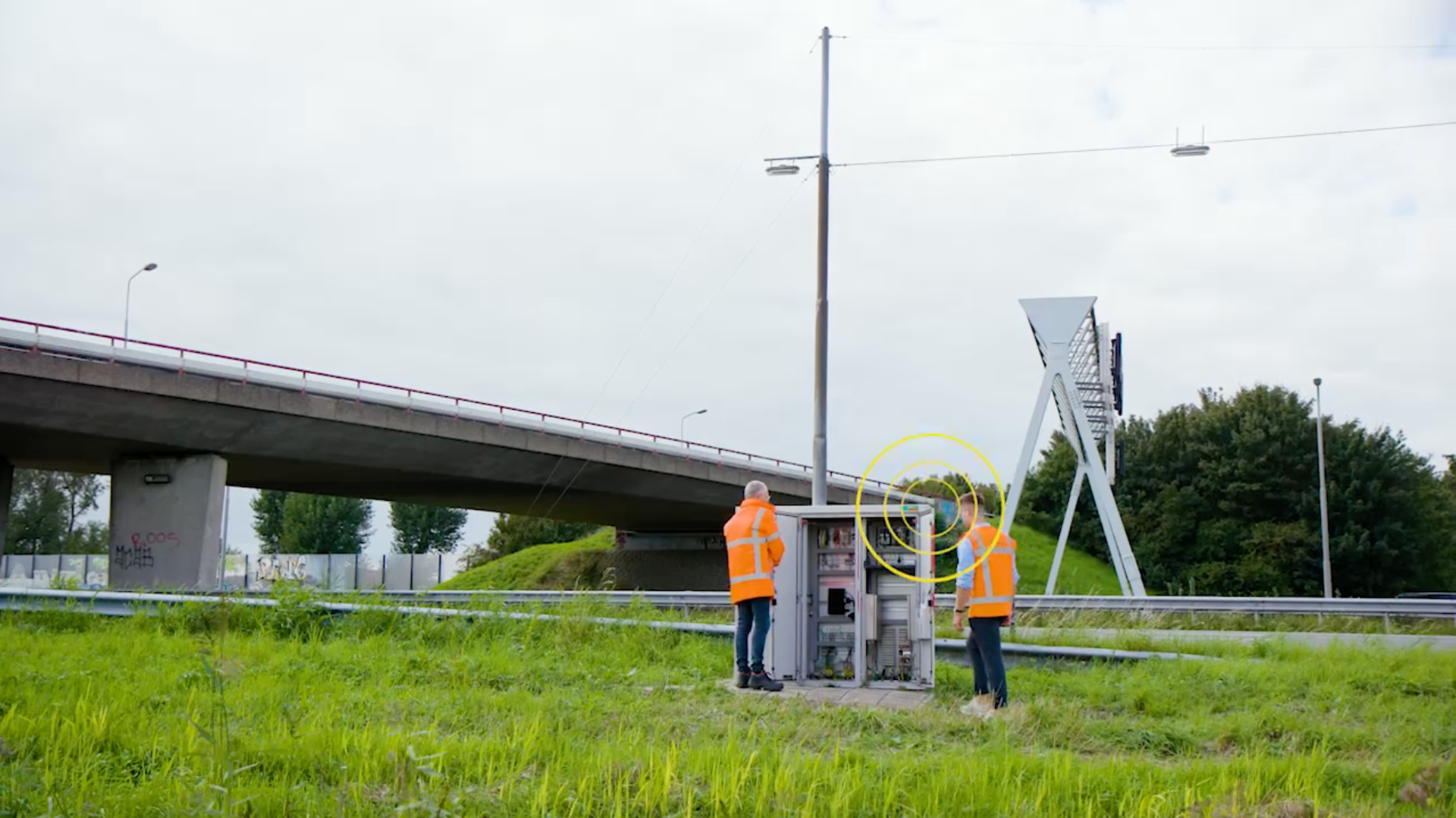 Twee medewerkers in veiligheidshesje bij meetapparatuur langs de snelweg naast een viaduct.