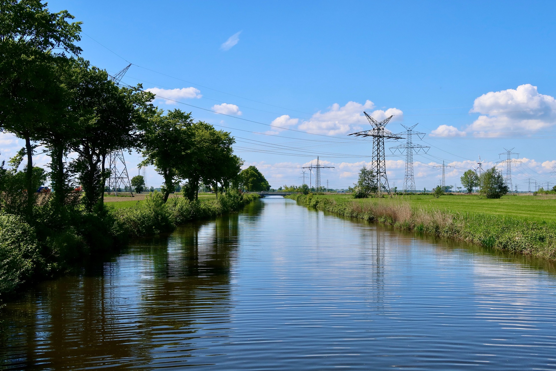 Een vredig zomerlandschap met een kanaal, elektriciteitskabels en weelderig groen onder een blauwe lucht.