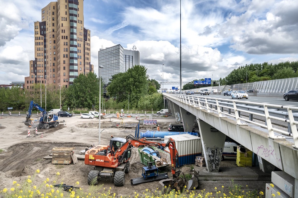 Aanleg van de nieuwe N200 Admiraal de Ruijterweg in Amsterdam, een bouwplaats met graafmachines, daarboven langs een brug en op de achtergrond de skyline van Amsterdam met kantorencomplexen.
